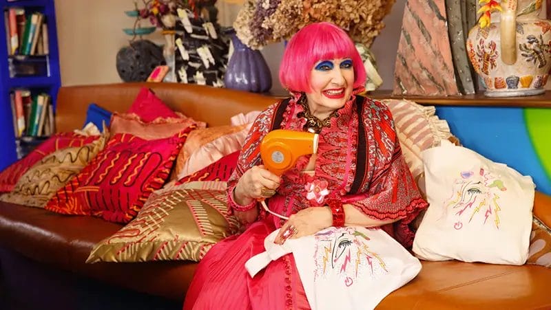Fashion designer Zandra Rhodes (a light skin tone woman with bright pink hair) sitting on a large sofa with her tote bag she designed to hold waste electricals