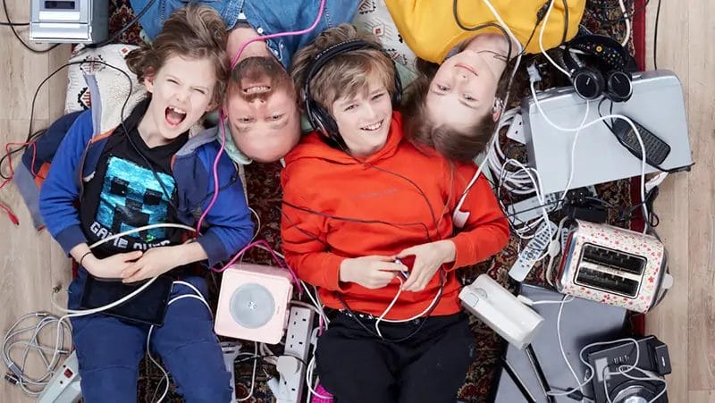 A light skinned family of a father and three children lying on the floor surrounded by hundreds of household electrical devices