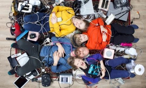 A pale skinned family of a father and three children lying on the floor surrounded by hundreds of household electrical devices