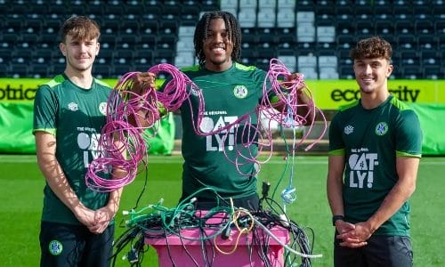 two pale skinned men and one dark skinned man in green football strip standing in front of a pink plastic wheelie bin full of cables. One of the men is holding up handfuls of cables and smiling