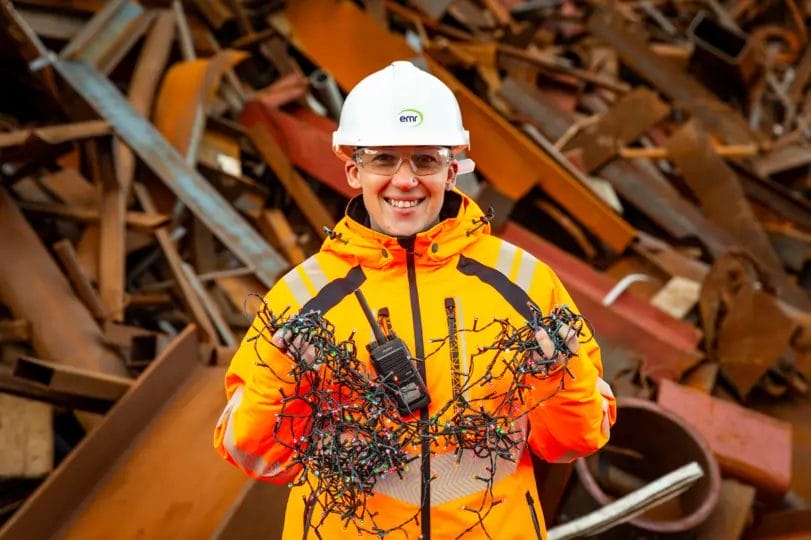 Sammy from EMR in a high-vis coat, hard hat and goggles holding up Xmas fairy lights and smiling at the camera